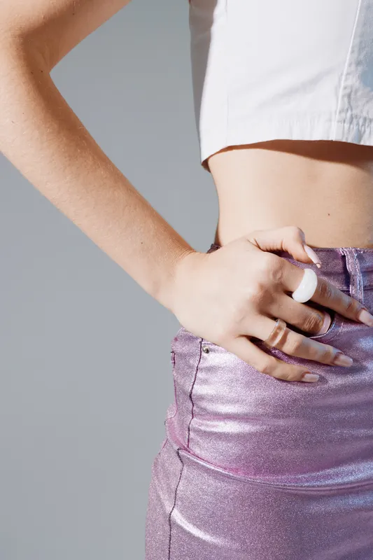 Close-up of a woman beauty fashion wearing a shiny purple skirt and white top. Her hand with a white ring rests on her waist. Studio shot on grey background.