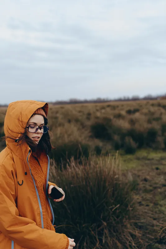 Woman outdoors field autumn jacket hood glasses lifestyle portrait nature, young woman in orange hooded jacket and glasses in grassy field, countryside hills, soft daylight, calm