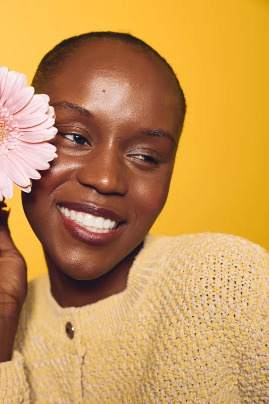 Close-up of happy black woman with dark skin holding pink flower near face, smiling in yellow knitted sweater on yellow background. Concept of beauty, joy, and natural skincare.