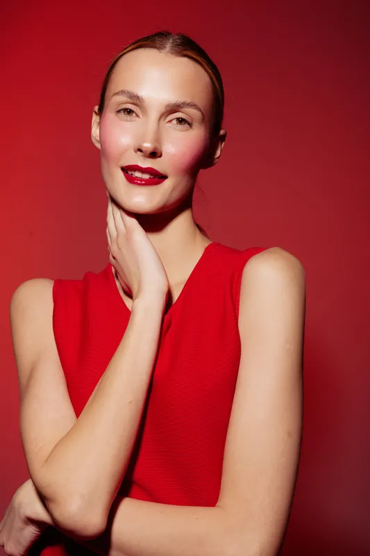 Young woman with red lipstick and red sleeveless top poses against red background. She touches her neck and smiles softly. Concept of beauty, fashion, makeup, and confident female model.