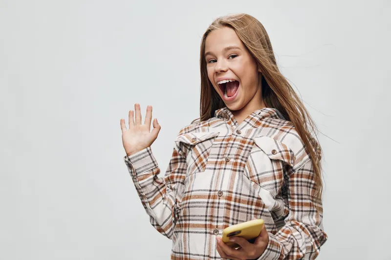 Teenage girl with long hair smiling and waving hand while holding yellow smartphone on light background. Casual checkered shirt and joyful expression for communication.