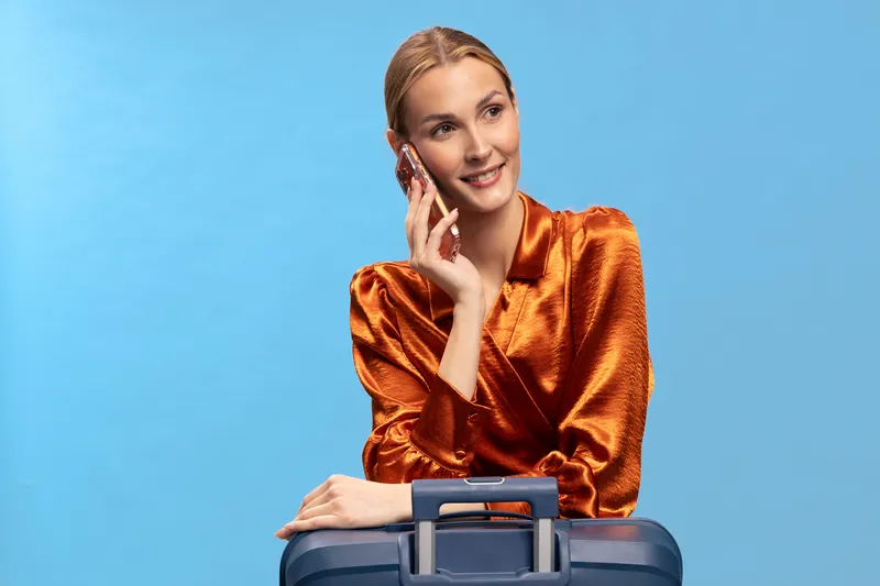 Young woman in orange satin blouse talks on smartphone and leans on blue suitcase against blue background. Concept of travel, communication, and business trip preparation.