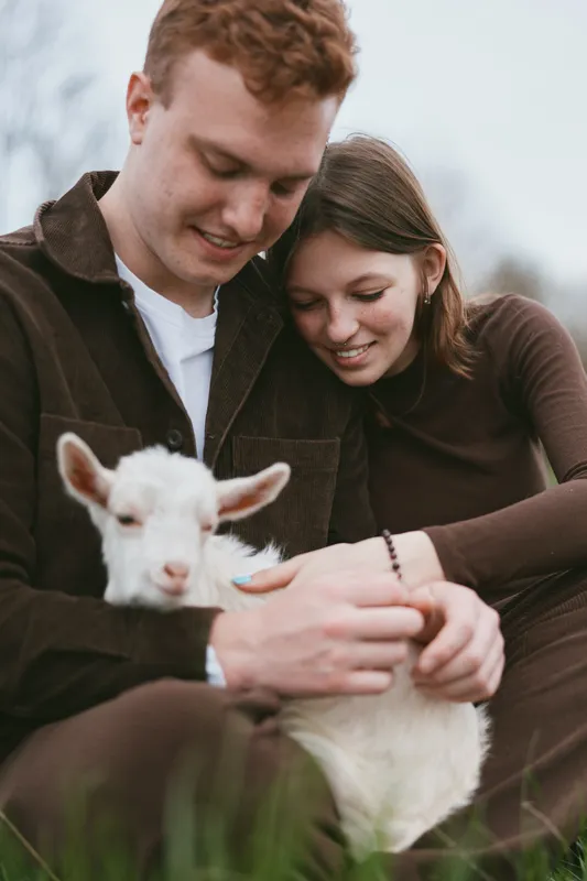 Couple shares a tender moment with a goat kid outdoors, enjoying rustic charm, calm smiles, and gentle interaction on a sunlit grassy field