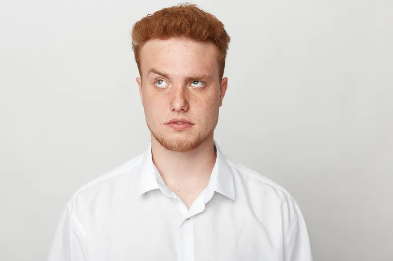 Young redhead man in white shirt looks up with thoughtful expression on light background. Concept of contemplation, decision making, and introspection in casual style.