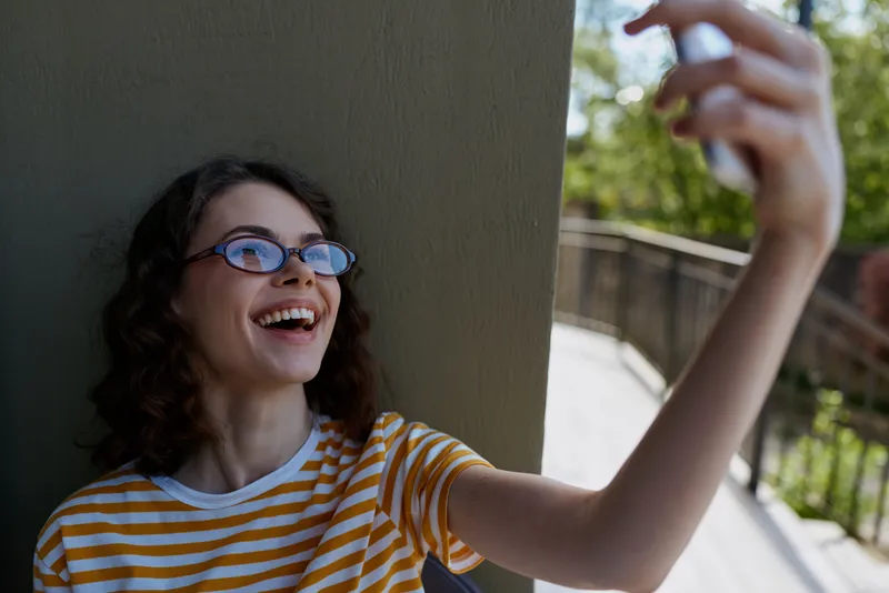 Smiling woman with curly hair and glasses taking a selfie on a balcony, wearing a striped shirt. Casual lifestyle moment outdoors, candid and cheerful. natural light adds warmth soft.