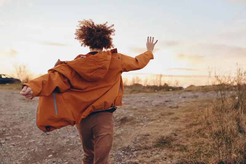 Woman lifestyle running outdoors autumn jacket in open field, back view, curly hair blowing, joyful motion, light dust in air, warm sunset landscape, candid lifestyle portrait at dusk
