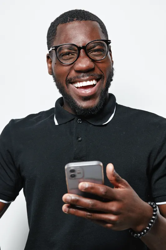 African American man with dark skin wearing glasses and black polo shirt smiling while holding smartphone in hand. Happy male using mobile phone for communication and technology.