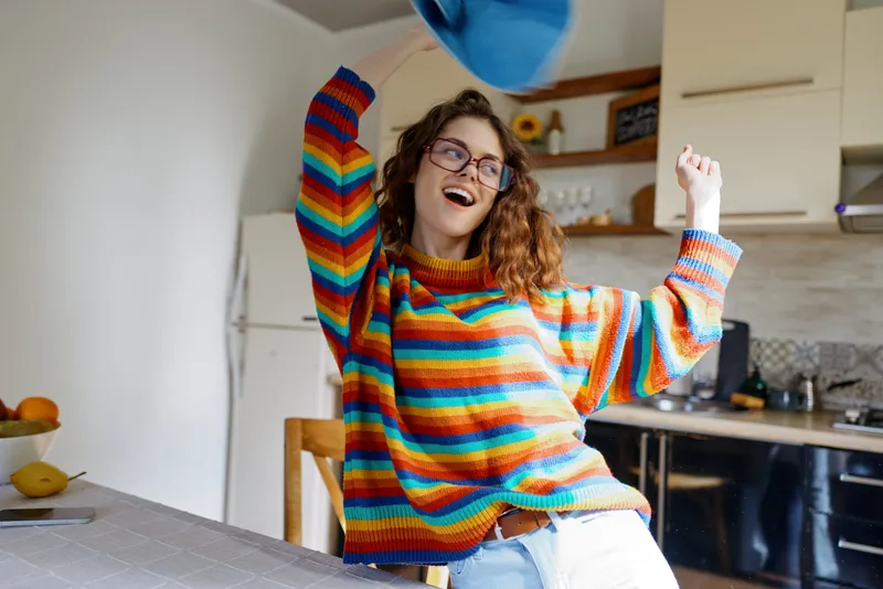 Cheerful woman in a colorful knit sweater dances in a bright kitchen, throwing arms up in joy, sunlight highlighting her smile and casual homey vibe during a candid lifestyle moment