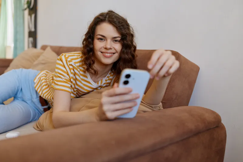A young woman lounges on a brown sofa at home, taking a casual selfie with her phone and smiling.