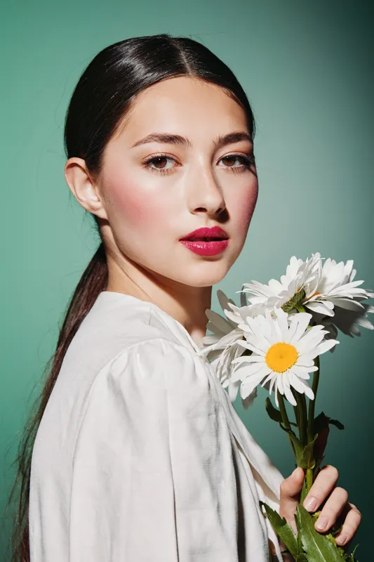 Young woman with bright makeup and glossy lips poses in white blouse holding daisies against green background. Fashion portrait emphasizing beauty, elegance, and floral aesthetic.