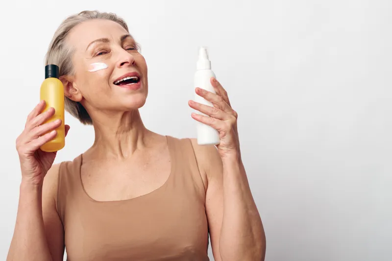 Smiling mature woman with skincare cream on face holding lotion and spray bottles in hands, enjoying beauty routine and self-care on white background with natural light.
