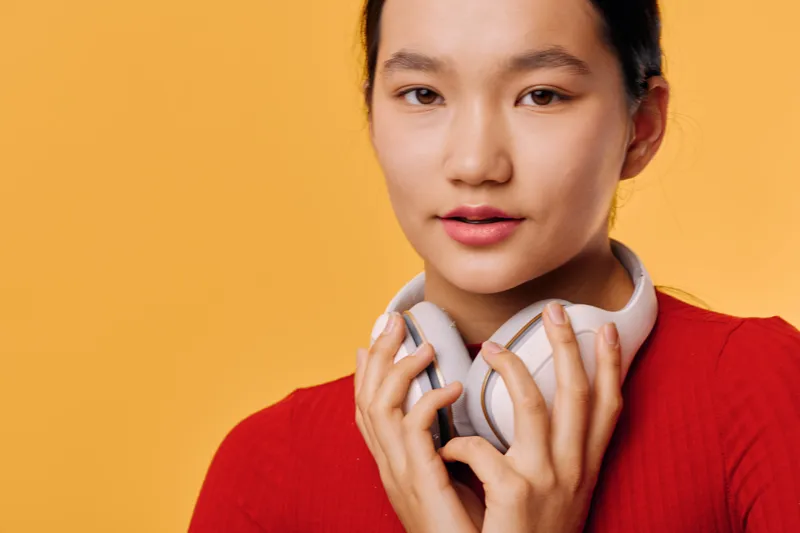 Young woman wearing white headphones around neck, looking at camera with slight smile against orange background. Female person in red top with casual style and technology accessory.
