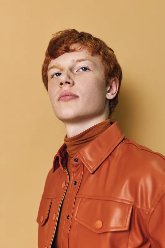 Portrait of young man with red hair wearing orange leather jacket and turtleneck looking confident on beige background in studio for fashion and style concept.