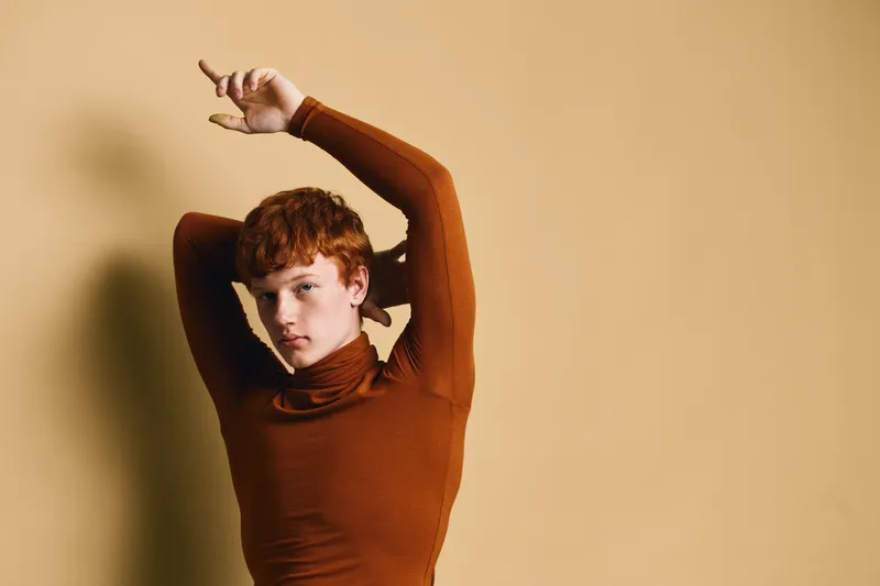 Young woman with short red hair wearing brown turtleneck posing against light brown background, portrait with raised arms for fashion, style, beauty and confidence concept.
