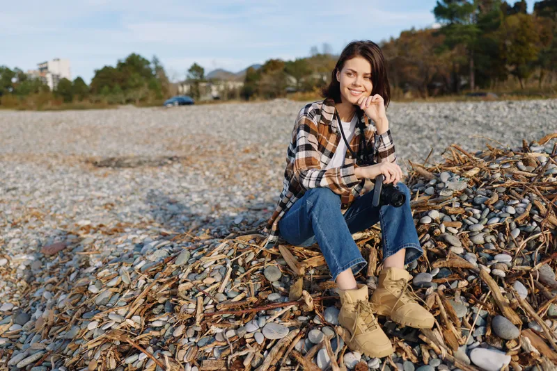 Woman on beach sits among rocks and pebbles in casual autumn attire, smiling softly under natural light, capturing a serene outdoor moment by the shoreline.