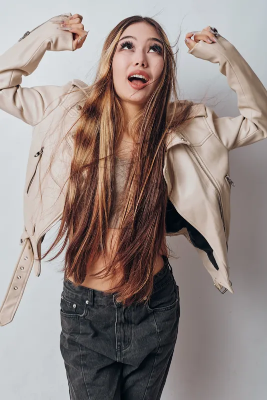Young woman with long hair wearing light jacket and black jeans posing with arms raised and looking up on gray background. Casual fashion style and confident expression in studio.