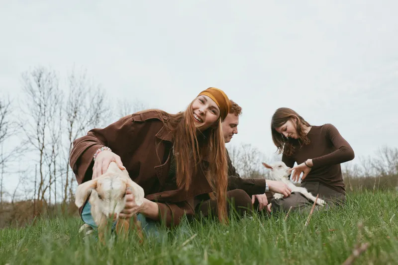 Young woman with long hair smiles while holding a baby goat in a grassy field with a man and woman playing with another goat in the background on a cloudy day.