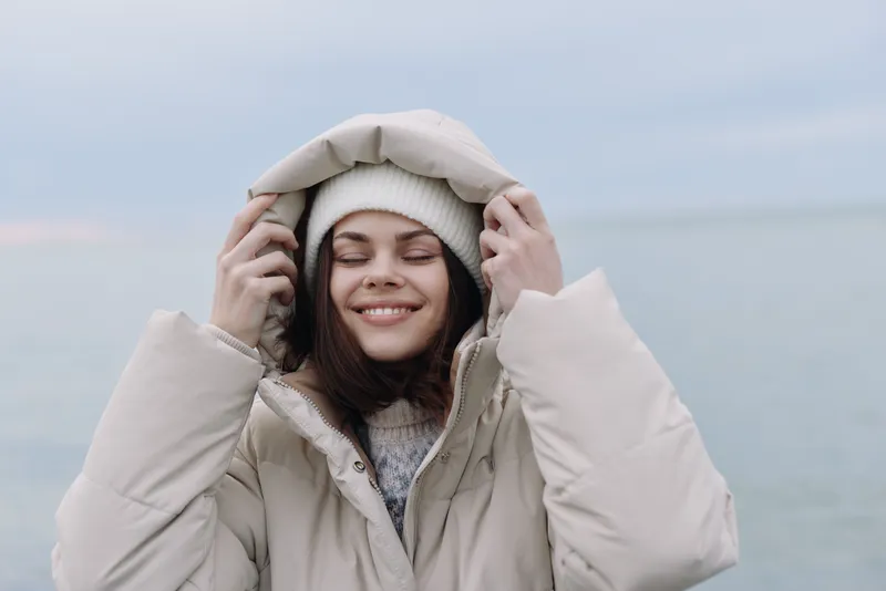 Girl in a warm coat and beanie smiling softly by the seaside, winter outfit and cozy mood capturing a quiet outdoor moment