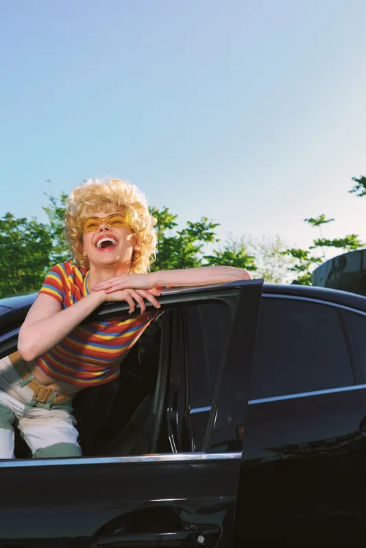 Joyful woman leans from a car window, laughing with a retro hairstyle and striped shirt, bright outdoor scene, cinematic color, casual lifestyle moment, playful mood.