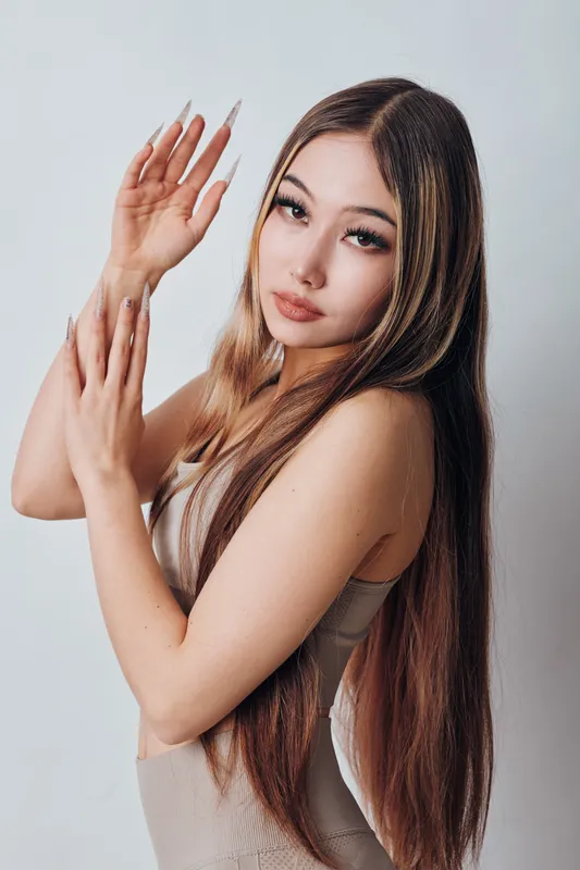 Young woman with long straight hair posing with raised arms against white background looking at camera with calm expression in studio portrait.