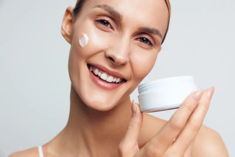 A smiling woman with clear skin applies white cream to her cheek while holding an open jar of moisturizer. The close-up portrait shows skincare, beauty, and healthy lifestyle concept.
