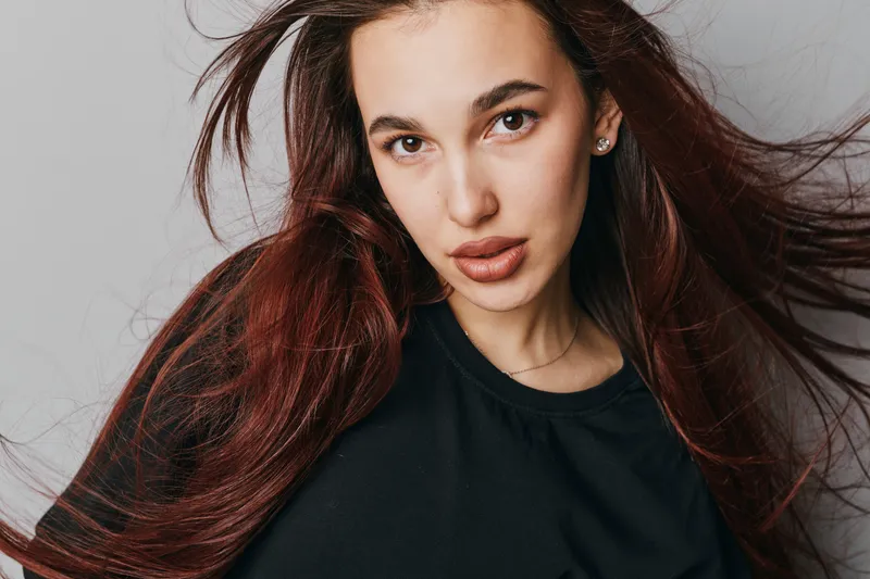 Woman with long brown hair and natural makeup posing indoors against gray background. Female model wearing black shirt with confident expression and flowing hair in studio portrait.