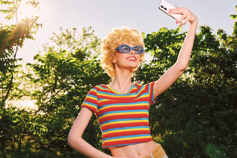 Smiling woman with curly hair posing outdoors, wearing a striped crop top and sunglasses, taking a selfie in bright sunlight. Retro film color vibe, vibrant lifestyle, carefree moment, in nature.