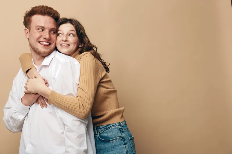 Young couple in casual clothes embraces and smiles, showing affection and happiness. The woman hugs the man from behind against a plain beige background. Concept of love and relationship.