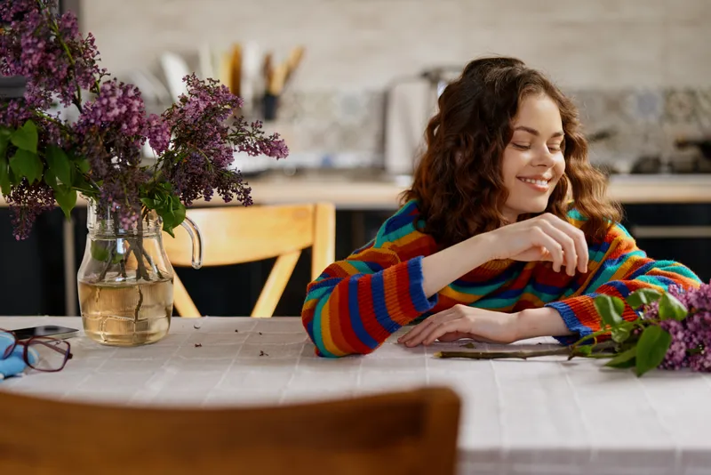 Woman sits at a home table with colorful striped sweater, smiling softly while arranging flowers, cozy kitchen setting, natural light, relaxed lifestyle moment, candid everyday mood