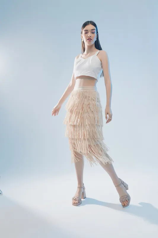 Young woman in a white crop top and beige fringed skirt poses confidently on a light background. Concept of woman beauty fashion and modern style with soft natural lighting.
