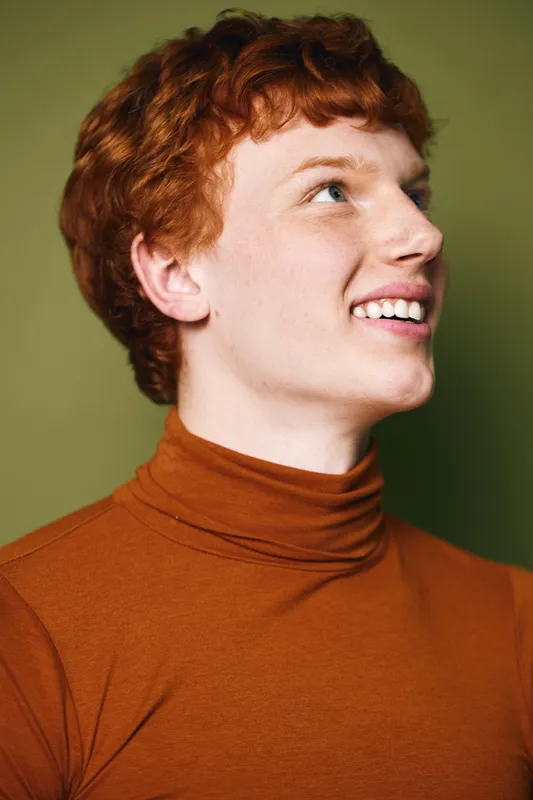 Portrait of young man with red hair wearing orange turtleneck smiling and looking up on plain background with natural light and positive expression.