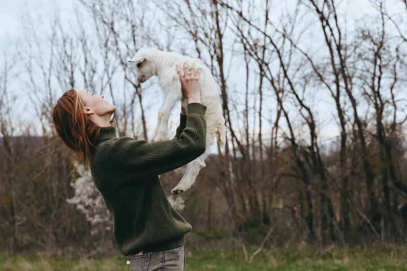 Smiling woman lifting a goat kid in an outdoor scene, wearing a green sweater. A candid lifestyle moment that highlights nature, happiness, affection, and playful bonding with animals in a serene