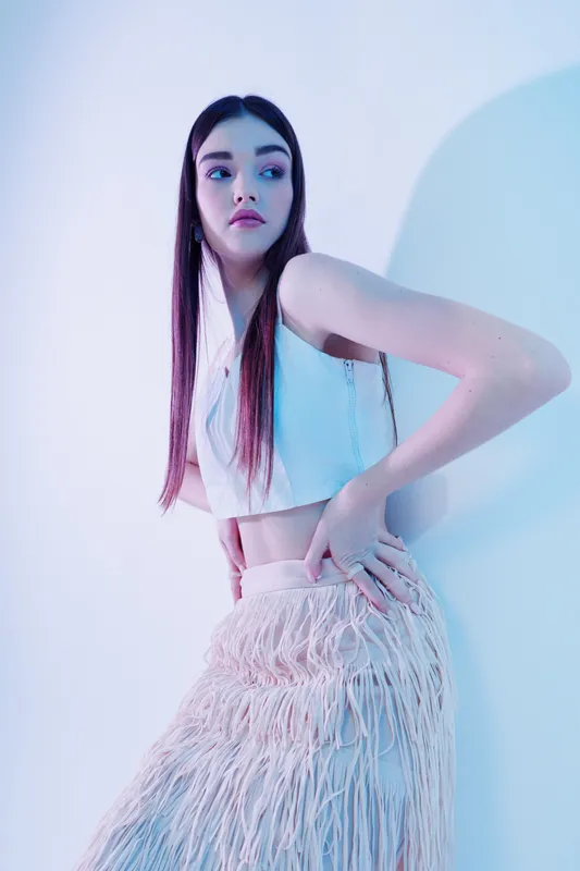 Young woman in fashionable white top and textured skirt poses against pastel background. Her long hair and confident expression highlight beauty and style. Concept for woman beauty fashion.