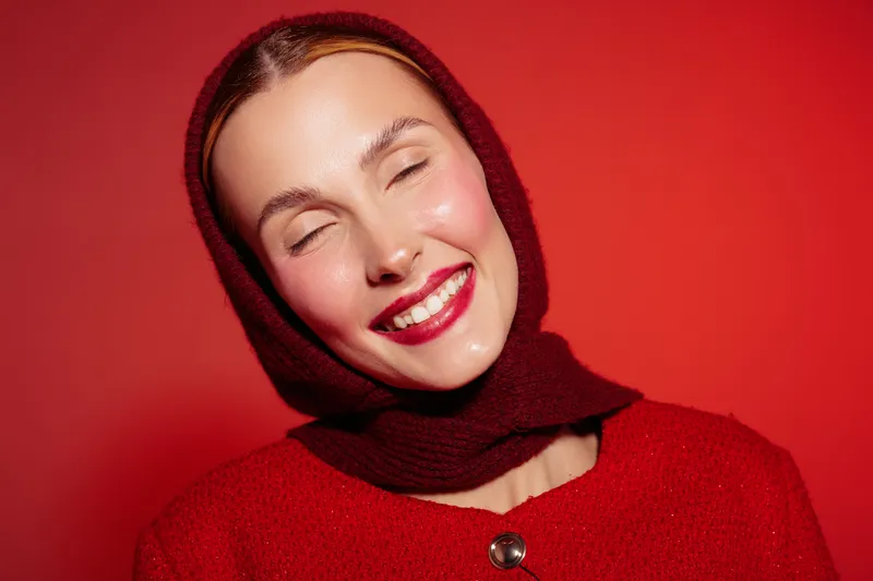 Smiling woman with closed eyes wearing red scarf and sweater on red background. Portrait of happy female model with bright lipstick and natural skin glow. Concept of joy and fashion.