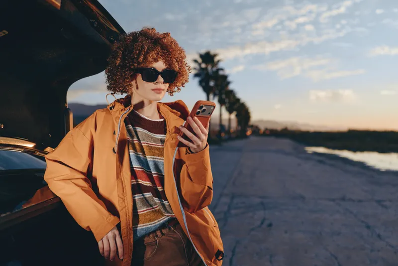 Woman lifestyle scene, curly haired woman in sunglasses holds smartphone by car on street at sunset, wearing orange jacket, near beach road at golden hour, relaxed smile, outdoor travel mood.