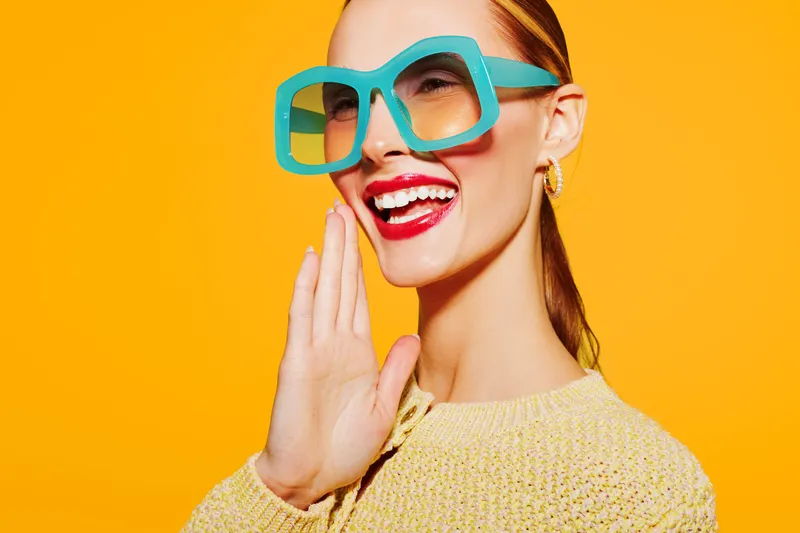 Smiling woman with bright red lipstick and large turquoise glasses laughs joyfully on yellow background. Concept of happiness, fashion, and vibrant lifestyle in studio lighting.