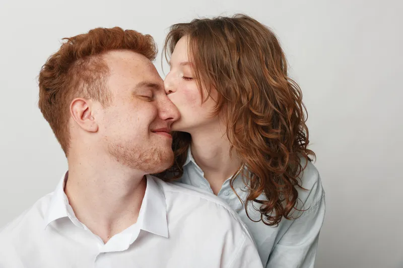 Young woman with curly hair kisses smiling man with red hair on cheek. Couple shows affection and love in casual clothes against plain light background. Concept of romance and intimacy.