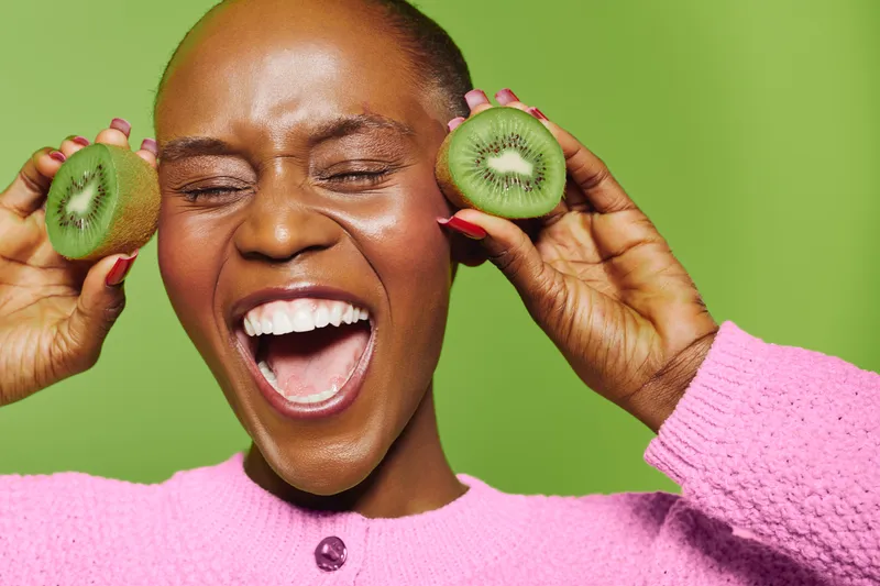Joyful black woman with dark skin laughing and holding two halves of kiwi fruit near her face. Bright green background and pink sweater create vibrant, fresh, and healthy concept image.
