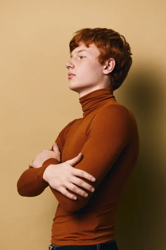 Young man with red hair wearing brown turtleneck stands with arms crossed, looking sideways against beige background. Casual fashion, confident pose, studio portrait with warm tones.