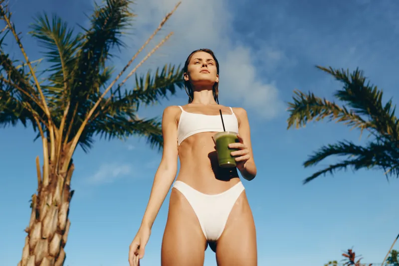 Young woman in a white bikini stands outdoors under a clear blue sky, holding a green smoothie cup, sunlit skin, palm trees framing the scene, conveying carefree summer mood and healthy lifestyle.