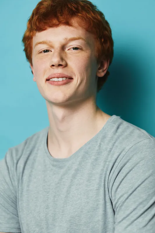 Young man with red hair and fair skin wearing gray shirt smiles confidently on blue background. Portrait of male with casual style and natural lighting for lifestyle or fashion.