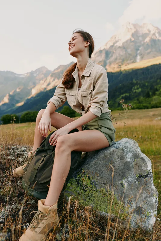 Young woman sits on a rock in a mountain meadow, wearing a beige shirt and green shorts, enjoying the fresh air, distant peaks, and a relaxed, carefree mood.
