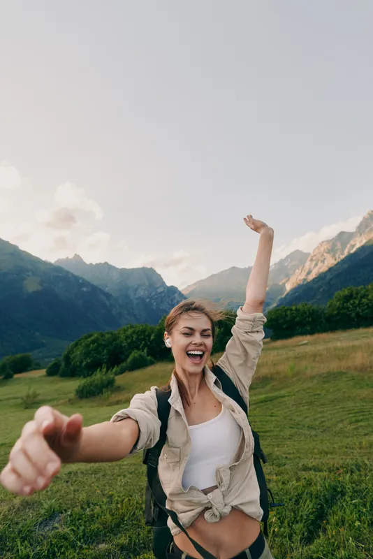 A joyful woman hikes through a green valley with distant mountains and a backpack, capturing a candid outdoors lifestyle moment of energy, freedom, and exploration.