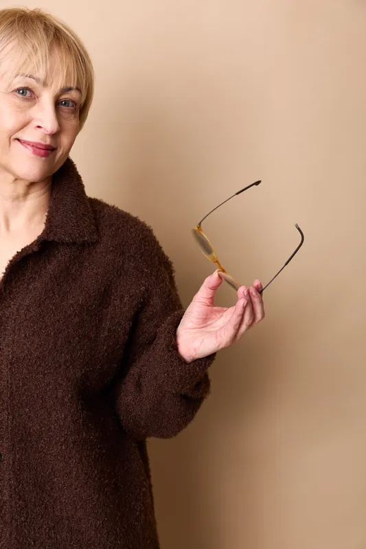 Senior woman in brown coat holding glasses in a studio portrait on beige background, relaxed smile, thoughtful mood, eyewear ready for daily life