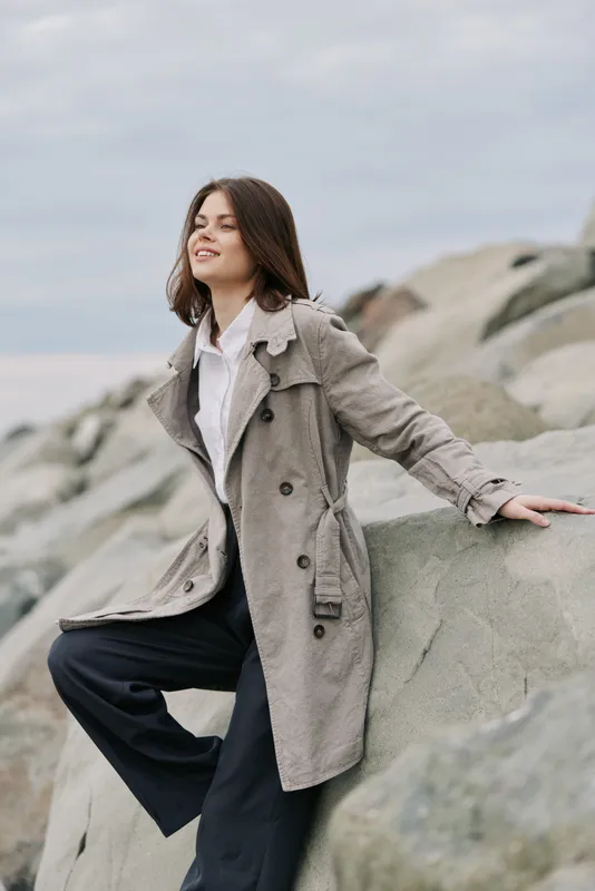 Young woman in a gray coat sits on large rocks outdoors, smiling and looking away. The cloudy sky and natural rocky background create a serene atmosphere.