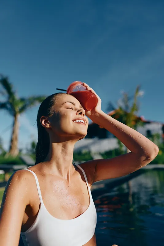 A young woman in a white swimsuit refreshes herself with a cold drink by the pool under a clear blue sky. Concept for summer lifestyle, relaxation, and wellness.
