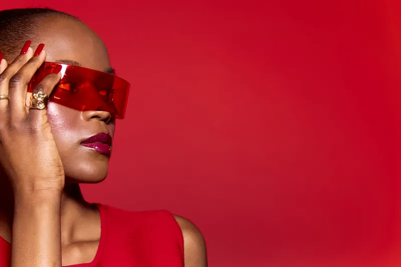 Stylish African American woman with dark skin wearing red sleeveless outfit and red futuristic sunglasses poses against red background. Concept of fashion, confidence, and modern style.