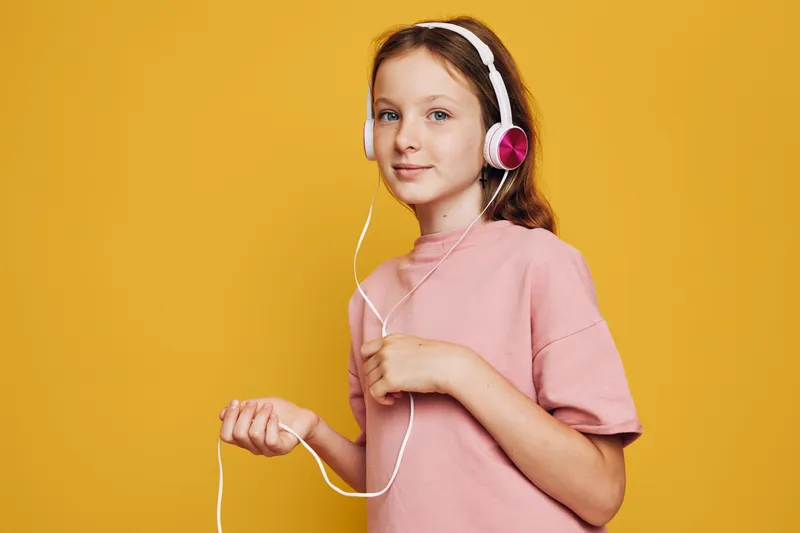 Young girl in pink shirt listens to music with white and pink headphones. She stands against yellow background with a relaxed and confident expression. Concept of youth and technology.