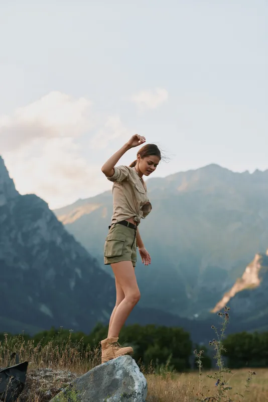 Young woman in casual outfit stands on a rock in a mountainous landscape during golden hour, enjoying nature and feeling free and confident.
