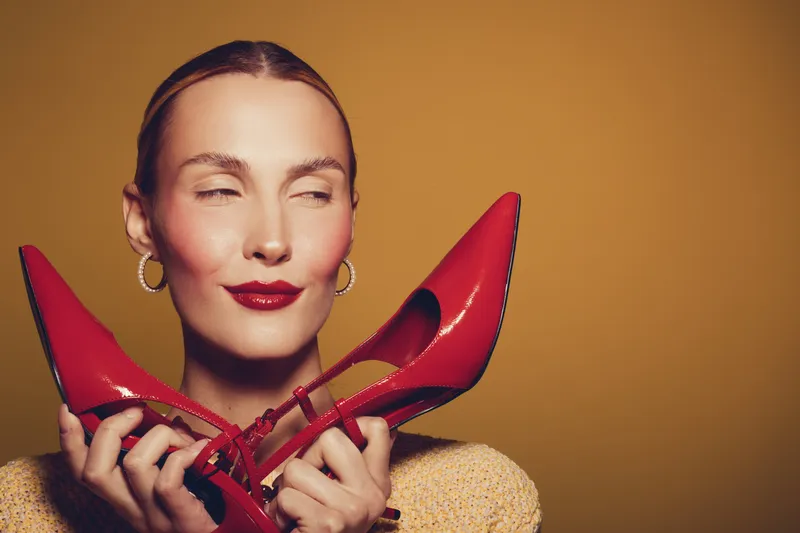 Woman with closed eyes holds red high heels near face, smiling softly. Warm studio lighting highlights her makeup and golden earrings on a plain brown background. Concept of fashion and style.